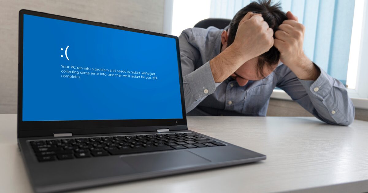 Um homem de camisa azul está sentado à mesa com as mãos na cabeça, aparentendo frustração. Na frente dele, há um laptop aberto exibindo a tela azul de erro do Windows, também conhecida como "tela azul da morte" (BSOD), com o ícone de um rosto triste ":(" e uma mensagem informando que o PC encontrou um problema e precisa reiniciar. O ambiente sugere um escritório, com uma janela coberta por persianas ao fundo. (falha tecnológica)
