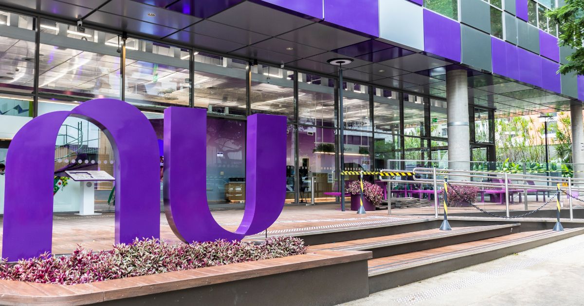 A imagem mostra a fachada de um edifício moderno, com amplas janelas de vidro e painéis externos na cor roxa e cinza. Em destaque, há uma grande escultura roxa em formato de "n" e "u" estilizados, que parece representar o logo do Nubank. Em frente à entrada do edifício, há uma rampa de acesso com corrimões metálicos e faixas amarelas e pretas de segurança. Vaso com plantas roxas e verdes decoram o espaço, harmonizando com o design moderno e colorido do local. Ao fundo, pode-se ver mais vegetação e uma área externa com bancos e mesas. (OXXO)