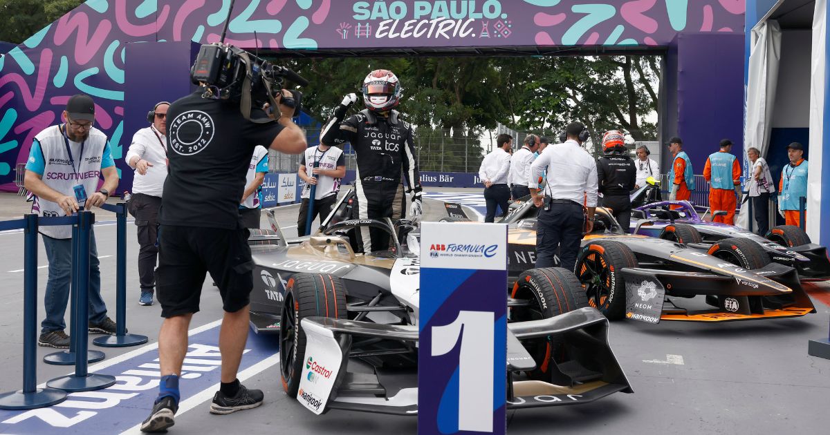 A imagem mostra um momento de celebração após uma corrida da Fórmula E em São Paulo. Em primeiro plano, está um carro de corrida com o número 1 destacado em um painel à frente, representando o vencedor. Um piloto em pé ao lado do carro, vestindo macacão e capacete, comemora com o braço levantado em gesto de vitória. Ao fundo, outros carros de corrida estão estacionados, com pessoas da equipe e oficiais circulando pelo local. O banner no alto exibe a frase “São Paulo Electric” com elementos gráficos coloridos. A cena inclui equipamentos de filmagem e fotógrafos capturando o momento, enquanto árvores e elementos urbanos compõem o ambiente externo, gemeos digitais