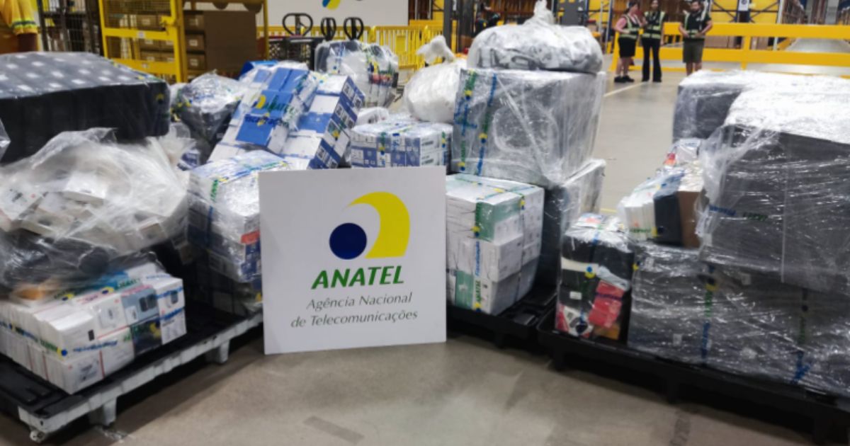 A imagem mostra diversos paletes envoltos em plástico transparente contendo caixas e equipamentos diversos. Em destaque, há um painel com o logótipo e o nome da ANATEL (Agência Nacional de Telecomunicações), que é o órgão regulador de telecomunicações no Brasil. Ao fundo, é possível ver uma área de armazenamento com pessoas em coletes de segurança próximas à cena. Os materiais parecem ser produtos apreendidos ou destinados à inspeção.