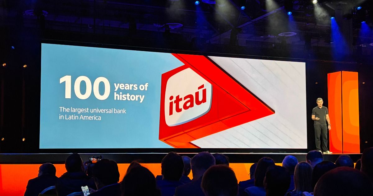 Na imagem, Ricardo Guerra, CIO do Itaú, está em um palco apresentando durante o re:Invent 2024, evento global da AWS. No telão ao fundo, destaca-se o logotipo do Itaú, com a mensagem: "100 years of history. The largest universal bank in Latin America". O ambiente conta com iluminação marcante, com tons de azul e laranja, enquanto o público, composto por executivos e profissionais de tecnologia, acompanha a apresentação em um auditório repleto. Ricardo está vestido de forma casual, reforçando a abordagem moderna e inovadora da palestra.