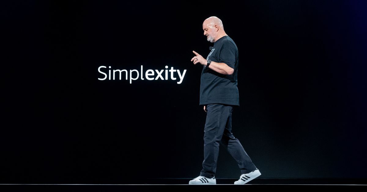Werner Vogels, CTO da Amazon, em uma apresentação no palco durante o re:Invent, o principal evento da Amazon Web Services (AWS). Ele caminha enquanto gesticula, ao lado da palavra "Simplexity" projetada em um fundo preto, representando o equilíbrio entre simplicidade e complexidade na inovação tecnológica. Com seu estilo casual, usando uma camiseta preta e tênis Adidas, Vogels reflete sua abordagem direta e prática ao discutir o futuro da tecnologia e seu impacto no mundo.