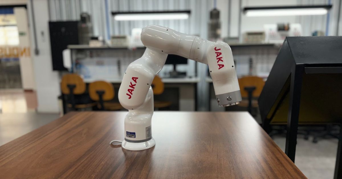 Robô colaborativo JAKA SCARA em um ambiente de laboratório, posicionado sobre uma mesa de madeira. Ao fundo, equipamentos, cadeiras e bancadas sugerem um cenário de pesquisa e desenvolvimento tecnológico (inatel, robo, scara, jaka)