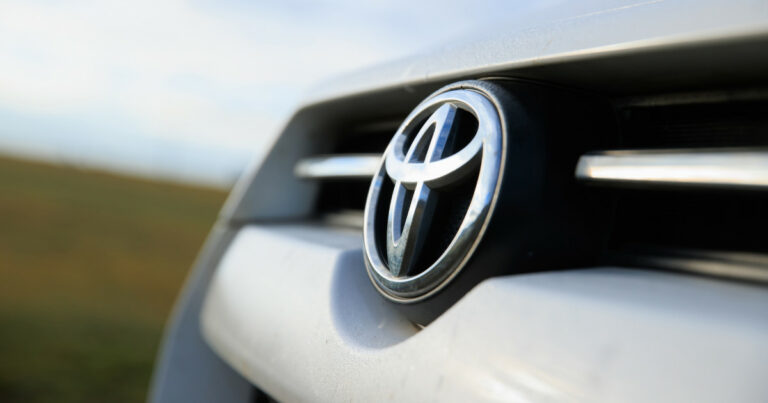 Close-up do emblema da Toyota na grade frontal de um veículo, destacando o logotipo prateado em alto relevo sobre fundo escuro. Ao fundo, desfocado, uma paisagem natural com céu claro