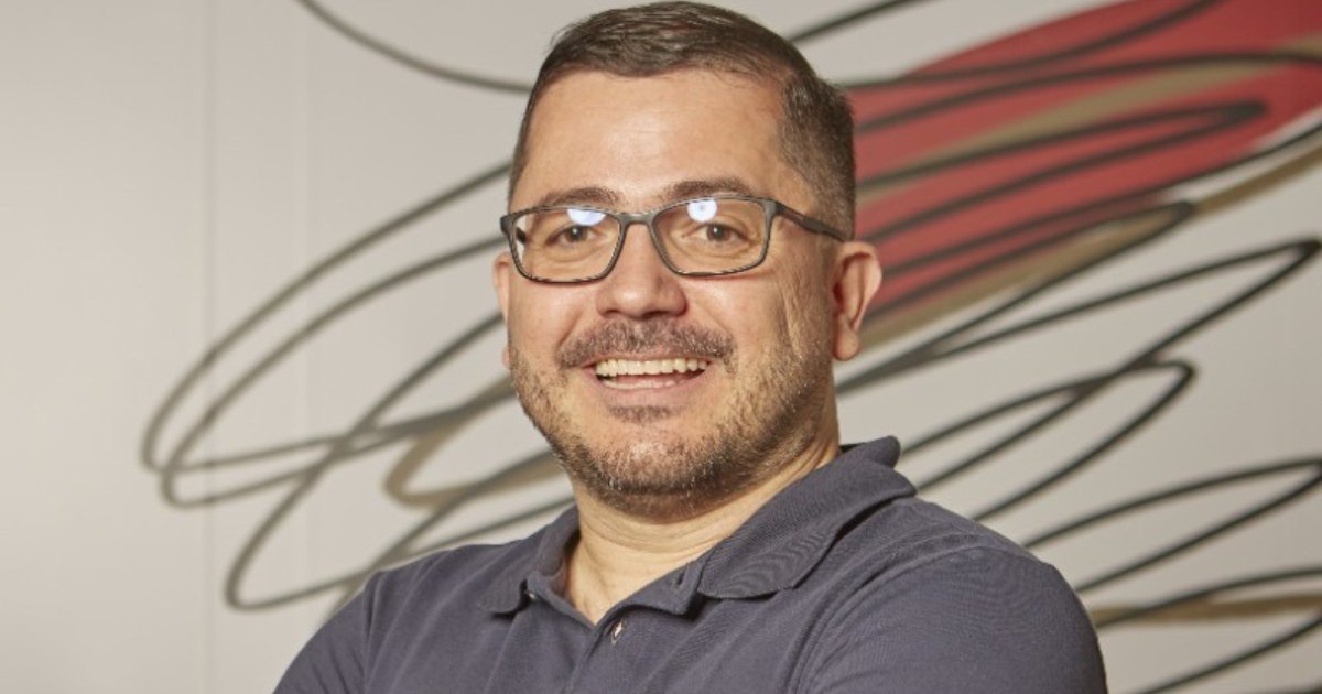 Foto de Everson Ramos, da Vórtx, sorrindo e posando com os braços cruzados. Ele usa óculos e uma camisa polo azul. O fundo apresenta um design abstrato com linhas em tons de vermelho e preto (projeto)