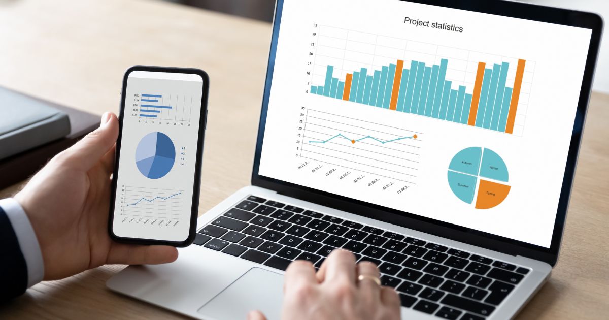 Uma pessoa segura um smartphone na mão esquerda enquanto utiliza um laptop com a mão direita, ambos exibindo gráficos e dados estatísticos. O smartphone apresenta gráficos de pizza, barras e uma linha em tons de azul. O laptop mostra uma tela com o título "Project statistics" (Estatísticas do projeto) e gráficos em colunas laranja e azul, um gráfico de linhas e um gráfico de pizza colorido. A cena ocorre em uma mesa de madeira clara, sugerindo um ambiente profissional ou de trabalho.