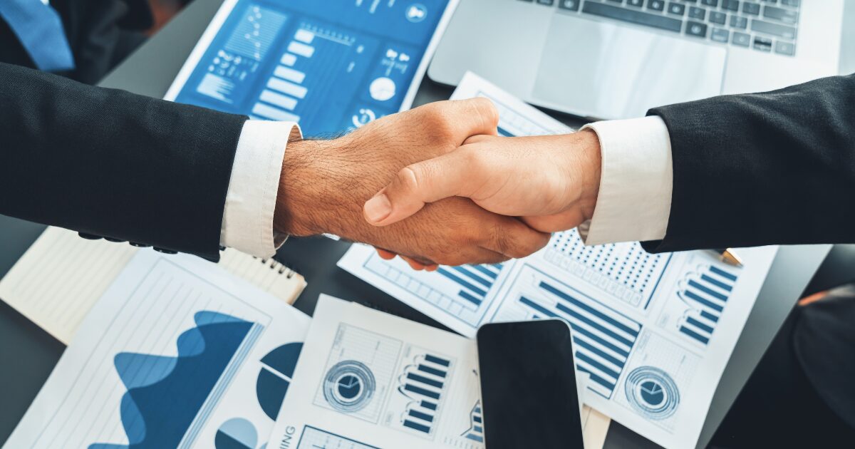 Duas pessoas em trajes formais apertando as mãos sobre uma mesa de escritório com documentos e gráficos financeiros espalhados. O cenário inclui um laptop, um celular e materiais com dados visuais, como gráficos de barras e linhas, simbolizando uma negociação ou acordo empresarial. A imagem transmite profissionalismo e colaboração no ambiente de negócios (colaboração, mundial, cooperação, negócio)