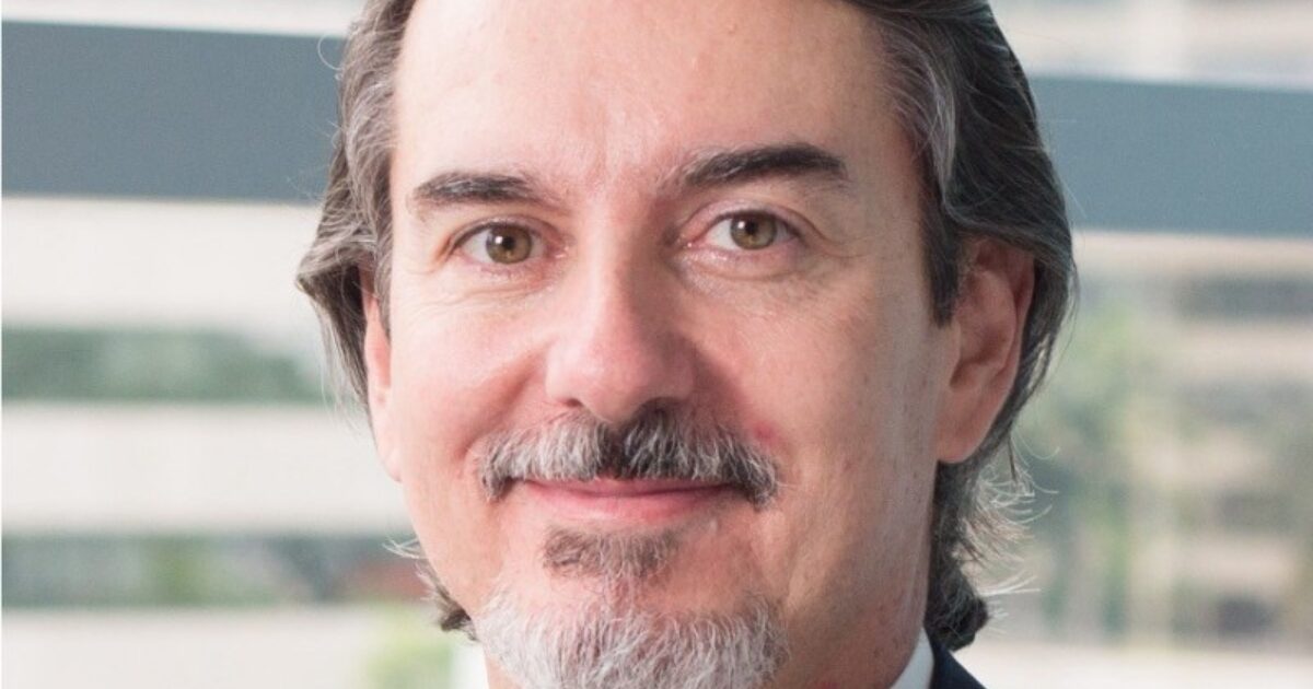 Fotografia de um homem com cabelos grisalhos, barba e bigode, sorrindo levemente. Ele está usando terno e gravata, com o fundo desfocado mostrando janelas e paisagem urbana. João Cerqueira, vice-presidente de Consultoria de Soluções para a América Latina na ServiceNow, fala sobre os agentes de IA