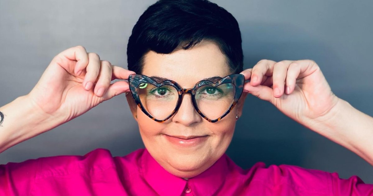 Mulher sorridente usando óculos em formato de coração com armação estampada. Ela veste uma camisa rosa vibrante e está ajustando os óculos com as mãos. O fundo é neutro e cinza, destacando o rosto e a expressão alegre da pessoa. (juliana nobrega, uniceub, Linkedin)