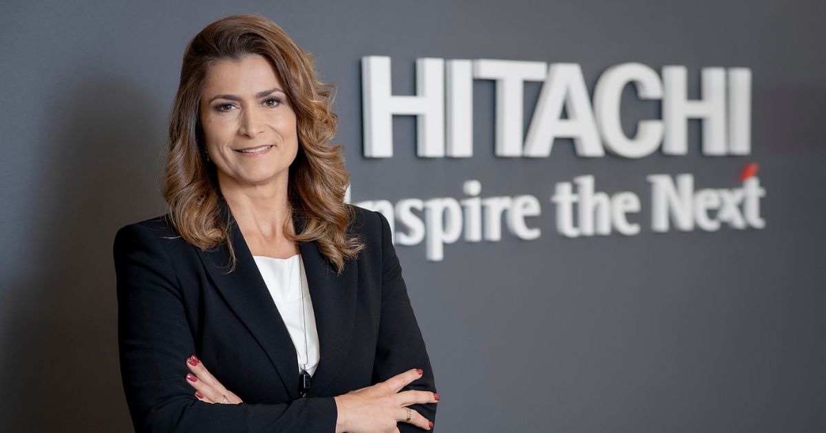 Andrea Fodor, CEO da Hitachi Vantara no Brasil, posa para uma foto corporativa em frente ao logotipo da empresa. Ela veste um blazer preto sobre uma blusa branca e mantém uma expressão confiante, com os braços cruzados. O fundo é um painel escuro com o slogan 'Inspire the Next' parcialmente visível, liderança
