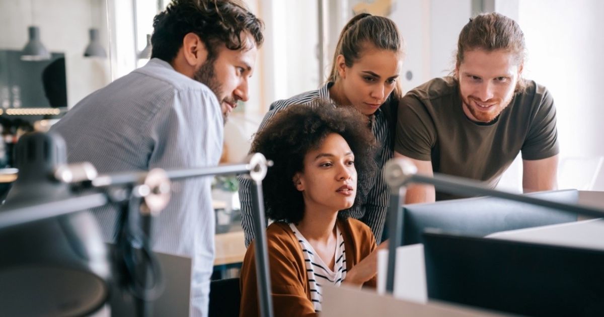 A imagem mostra um grupo de quatro jovens profissionais reunidos ao redor de um computador em um ambiente de escritório moderno. A mulher no centro, de cabelo cacheado, parece estar explicando algo na tela enquanto os outros três, um homem de barba, uma mulher de cabelo preso e outro homem de cabelo longo, observam atentamente. O ambiente tem um design contemporâneo, com luminárias pendentes e um espaço bem iluminado. A cena transmite colaboração e trabalho em equipe (positivo tecnologia, positivo S+)
