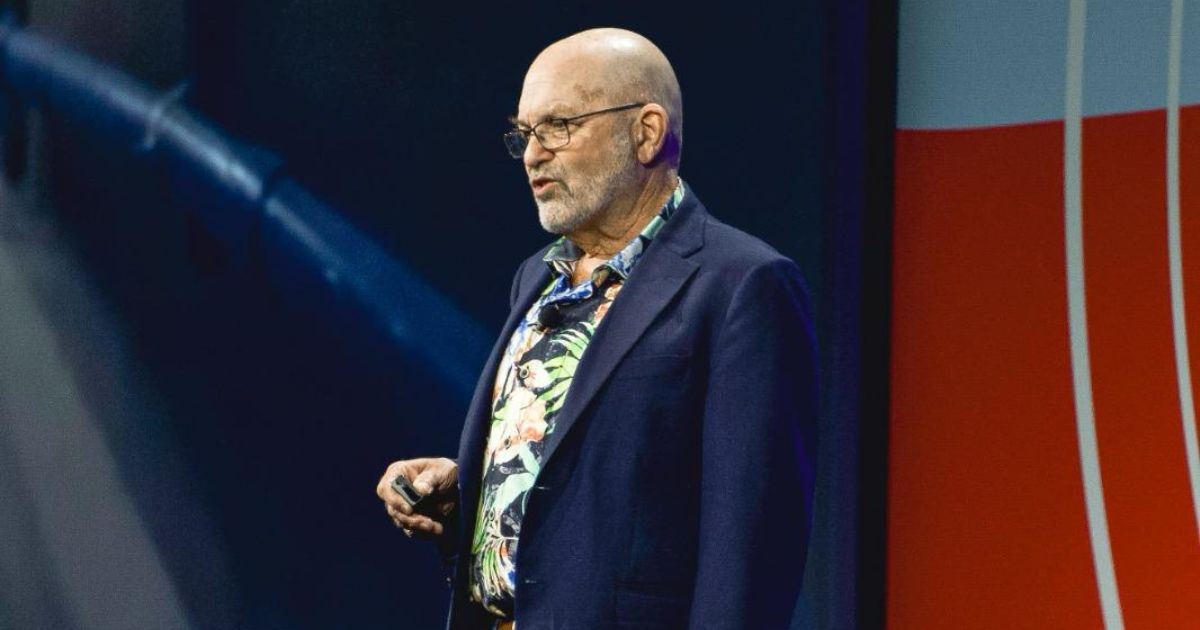 Marc Raibert, diretor-executivo do RAI Institute e fundador da Boston Dynamics, no palco do evento 3DExperience World 2025. Ele está à esquerda, vestindo um blazer escuro e camiseta, enquanto um grande telão ao fundo exibe o nome do evento "3DExperience World" com um design em tons de azul e vermelho. À direita, outro telão exibe a frase "I Love Robotics". A plateia, parcialmente visível no primeiro plano, assiste à apresentação em um ambiente iluminado com luzes cênicas