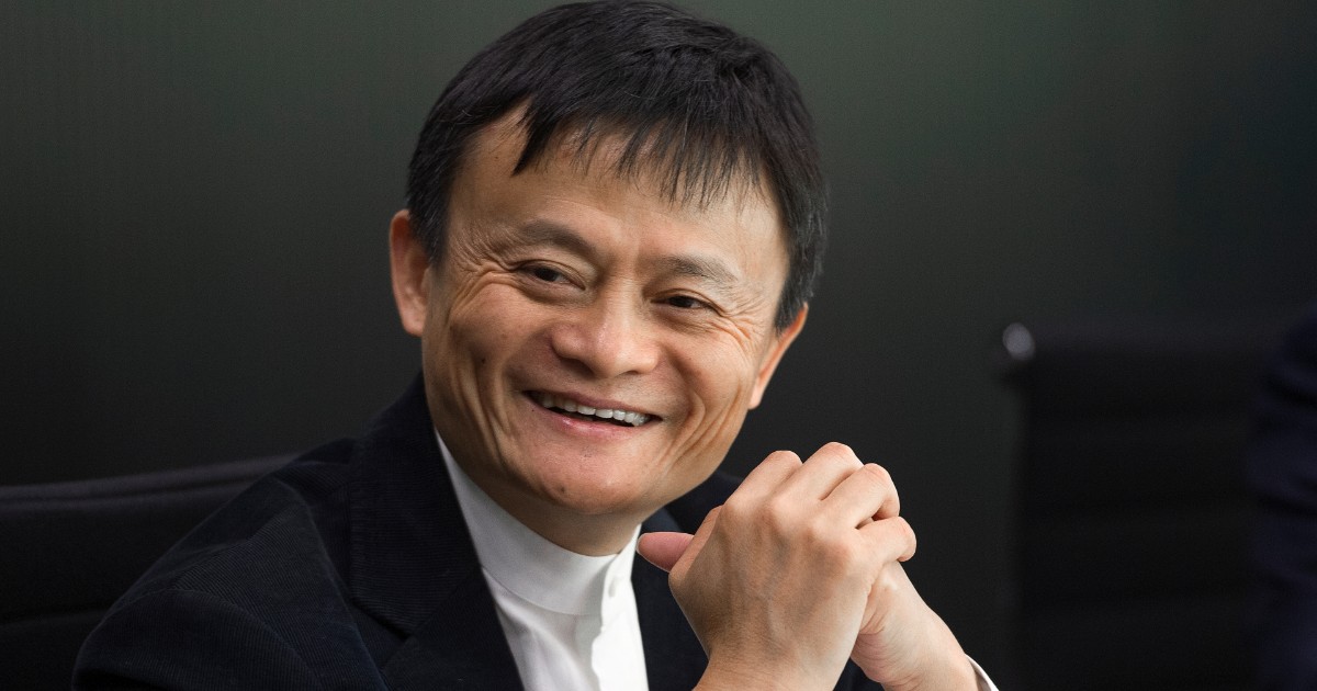 Esta é uma imagem de Jack Ma, cofundador do Alibaba, sorrindo em um ambiente corporativo. Ele veste um terno escuro com uma camisa branca e está sentado com as mãos entrelaçadas. O fundo escuro e desfocado sugere um ambiente de reunião ou conferência (china)
