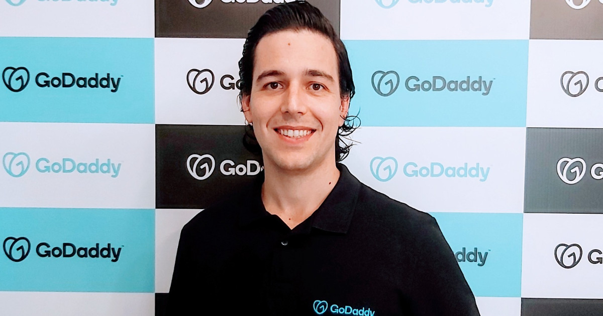 A imagem mostra um homem sorridente vestindo uma camisa polo preta com o logotipo da GoDaddy bordado no peito. Ele tem cabelo escuro penteado para trás e está posicionado em frente a um fundo padronizado com o logotipo da GoDaddy em tons de azul, preto e branco. O homem na imagem é Luiz D'Elboux, country manager da GoDaddy para o Brasil