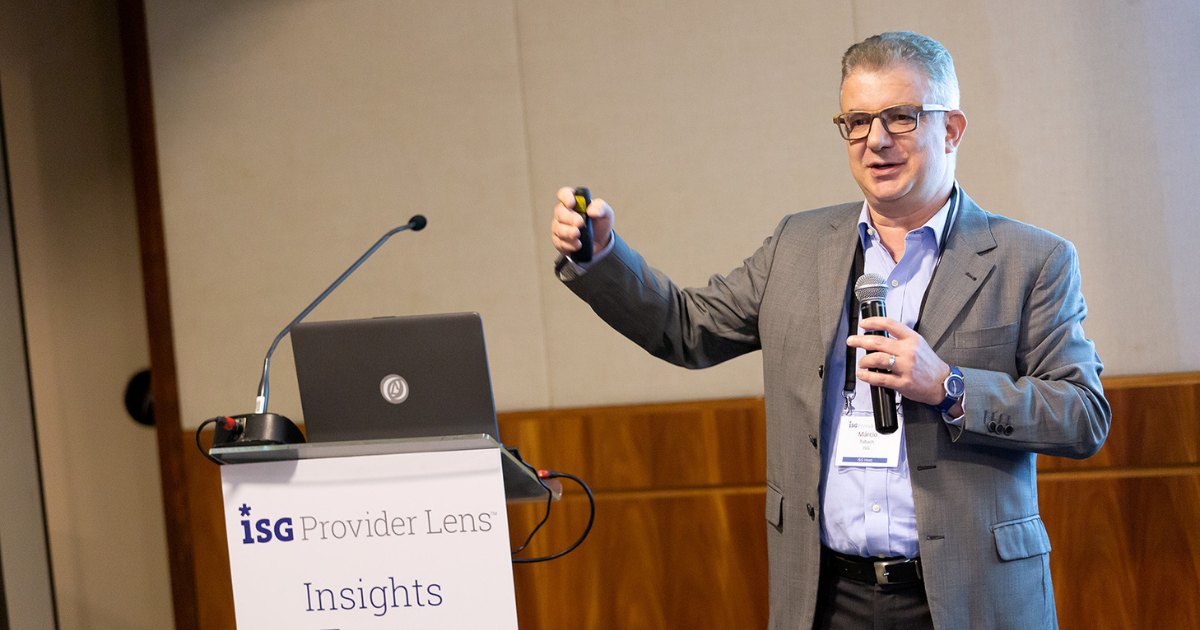 A imagem mostra Márcio Tabach, distinguished analyst da TGT ISG, durante uma apresentação no evento ISG Provider Lens Insights. Ele veste um terno cinza com camisa azul clara e usa óculos. Segura um microfone em uma mão e um controle de apresentação na outra, enquanto fala para o público. Ao fundo, há um púlpito com um notebook e o logotipo do evento, empresas