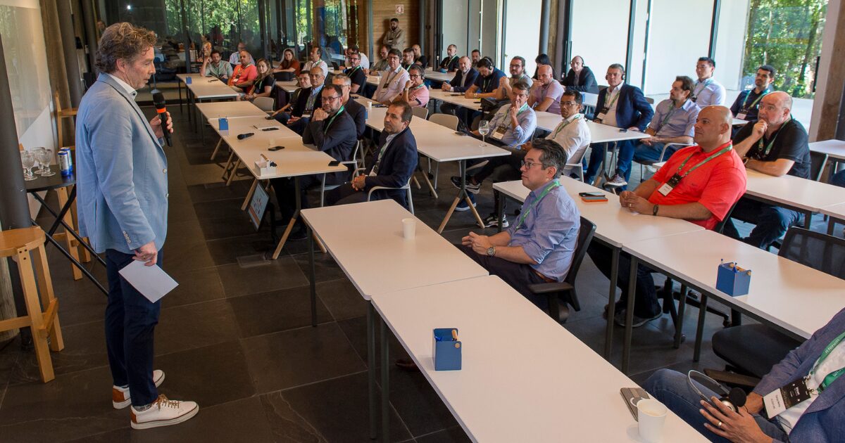 Gord Mawhinney, presidente das Américas e gerente geral dos EUA na Avanade, durante o IT Forum Na Mata. Ele está de pé à esquerda, vestindo um blazer azul-claro e segurando um microfone enquanto fala para um público atento. Os participantes estão sentados em mesas brancas, vestindo crachás e roupas formais, demonstrando interesse na apresentação. O ambiente possui janelas amplas, permitindo a entrada de luz natural e uma vista para a vegetação externa
