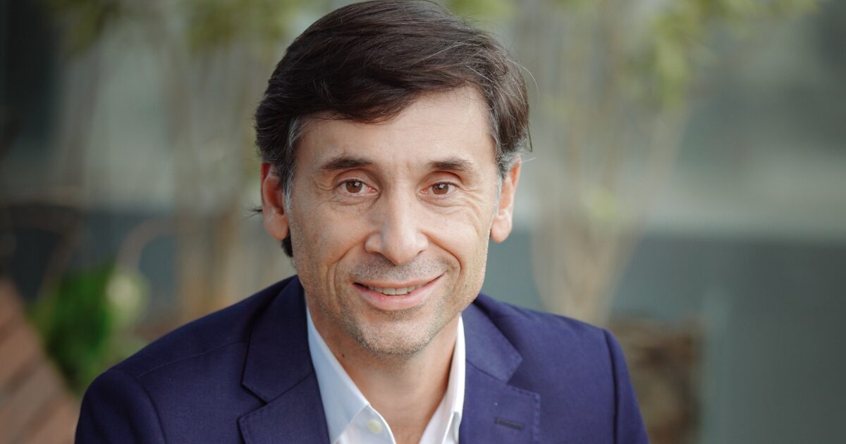Retrato de Willian Pimentel, vice-presidente da Freshworks na América Latina. Ele está sentado ao ar livre, vestindo um terno azul e camisa branca, sorrindo para a câmera. A imagem transmite uma atmosfera profissional e acessível. Software, IA