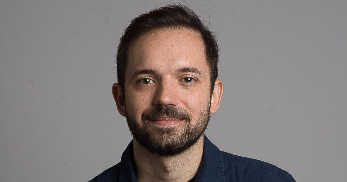 Retrato de Renato Pedigoni, CTO do Grupo Boticário. Ele tem barba curta e cabelo castanho escuro, penteado para o lado. Veste uma camisa azul e sorri levemente para a câmera. O fundo é neutro e cinza, com iluminação suave e equilibrada.