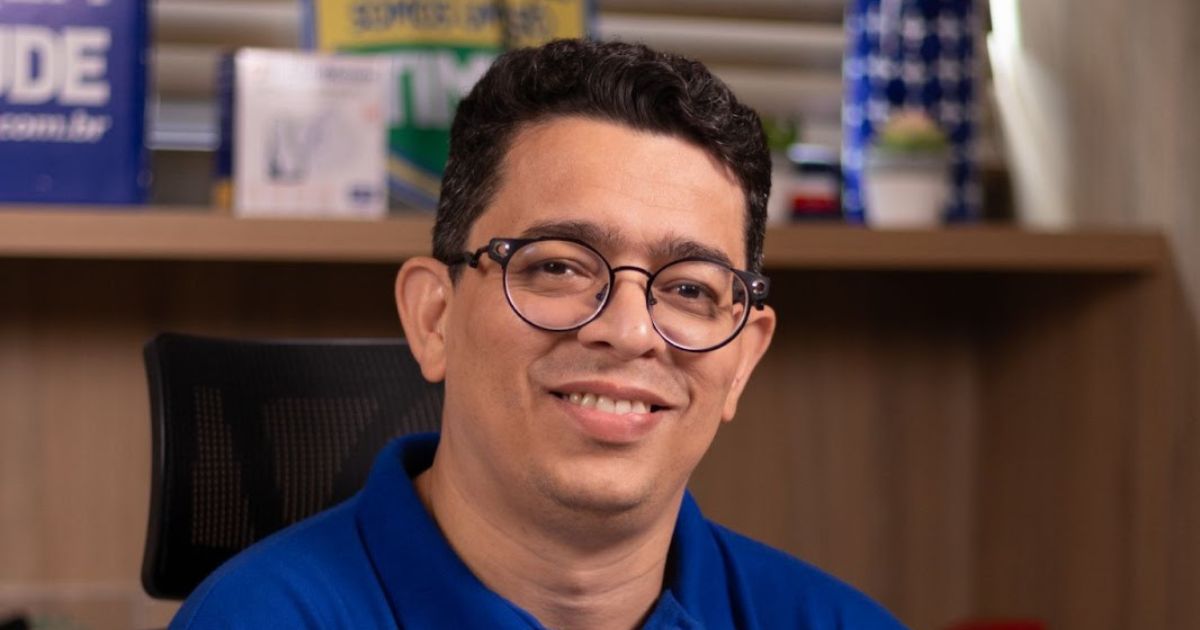 A imagem mostra Uderson Fermino, o novo diretor de TI da Pague Menos, sorrindo para a câmera. Ele usa óculos de aro redondo e uma camisa azul. O fundo da imagem apresenta um ambiente de escritório, com prateleiras contendo objetos decorativos e materiais corporativos.