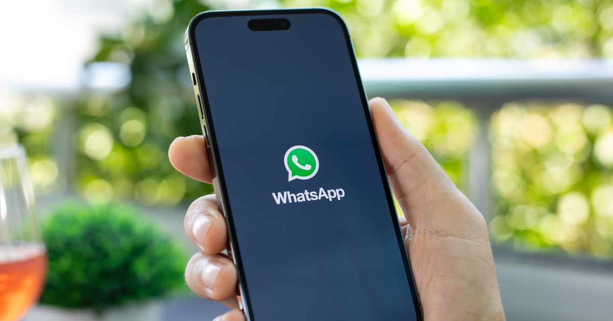A imagem mostra uma mão segurando um smartphone com a tela ligada, exibindo a tela de carregamento do aplicativo WhatsApp. O logotipo verde e branco do WhatsApp está centralizado na tela preta, com o nome do aplicativo logo abaixo. O fundo da imagem está levemente desfocado, mostrando um ambiente ao ar livre com plantas verdes e um copo com bebida alaranjada ao lado