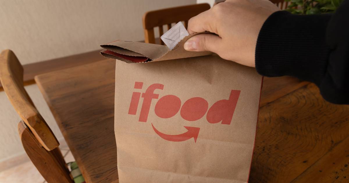 A imagem mostra uma mão abrindo uma sacola de papel pardo com o logotipo do iFood impresso em vermelho. A sacola está sobre uma mesa de madeira, e ao fundo há cadeiras de madeira e uma parede clara. Dentro da sacola, é possível ver um pequeno pacote de papel. A cena transmite a ideia de recebimento de um pedido de comida via delivery