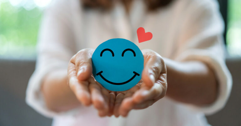 A imagem mostra uma pessoa segurando delicadamente um círculo azul com um rosto sorridente desenhado, representando positividade e bem-estar. Acima, há um pequeno coração vermelho, reforçando a ideia de cuidado e saúde mental. O fundo é desfocado, dando destaque às mãos e ao símbolo de felicidade