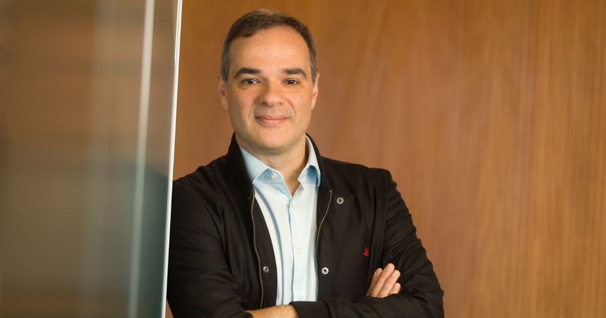 Felipe Ávila sorri levemente com os braços cruzados, usando uma camisa azul clara e jaqueta preta, em frente a uma parede de madeira.