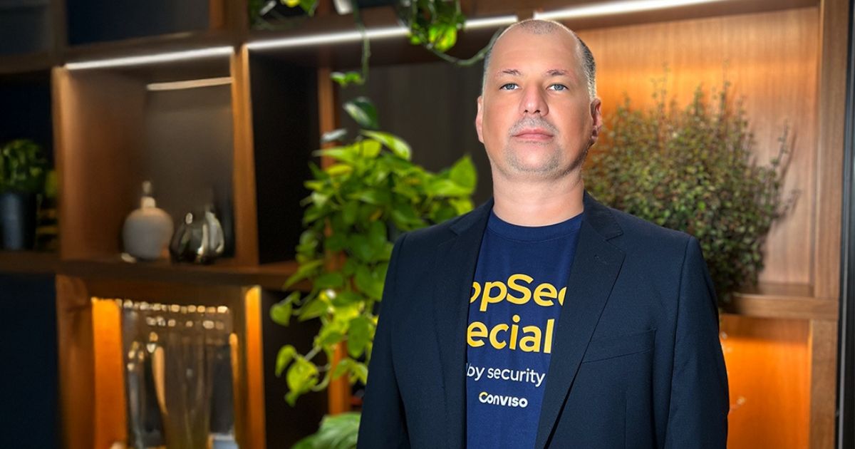 Um homem careca de pele clara, com um leve sorriso e expressão confiante, está posando para a foto. Ele veste uma t-shirt azul marinho com letras amarelas que dizem "AppSec Special", acompanhada por um blazer preto. Ao fundo, há uma elegante estante de madeira iluminada com diversas plantas e objetos decorativos, criando um ambiente sofisticado e moderno (conviso)
