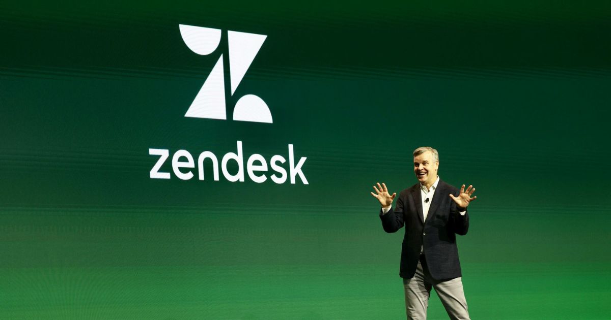 Um homem vestido com um blazer escuro, camisa branca e calças claras está no palco fazendo uma apresentação. Ele sorri e gesticula com as mãos abertas enquanto fala. Atrás dele, há um grande ecrã verde exibindo o logotipo da Zendesk, composto por um design geométrico branco e o nome da empresa em letras minúsculas. O ambiente sugere um evento corporativo ou conferência de tecnologia.