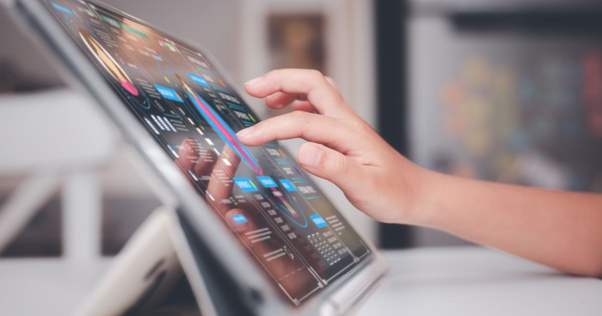 Mão interagindo com um tablet em um suporte, exibindo uma interface digital com gráficos, estatísticas e dados coloridos. A tela apresenta elementos visuais sofisticados, sugerindo uma análise de dados ou painel de controle digital. O fundo está desfocado, destacando o dispositivo e a interação do usuário (desafios)