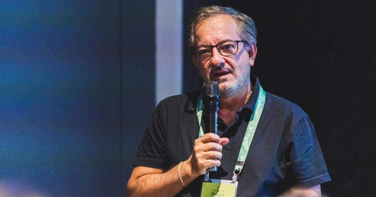 Homem de meia-idade, com barba grisalha e óculos, segura um microfone enquanto fala durante um evento. Ele usa uma camisa polo preta e um crachá verde suspenso no pescoço com a inscrição "Itaqui" e "IT Forum". O fundo da imagem é um painel azul desfocado. Nelson Campelo, CEO da Atos para a América Latina durante o Jornada Itaqui