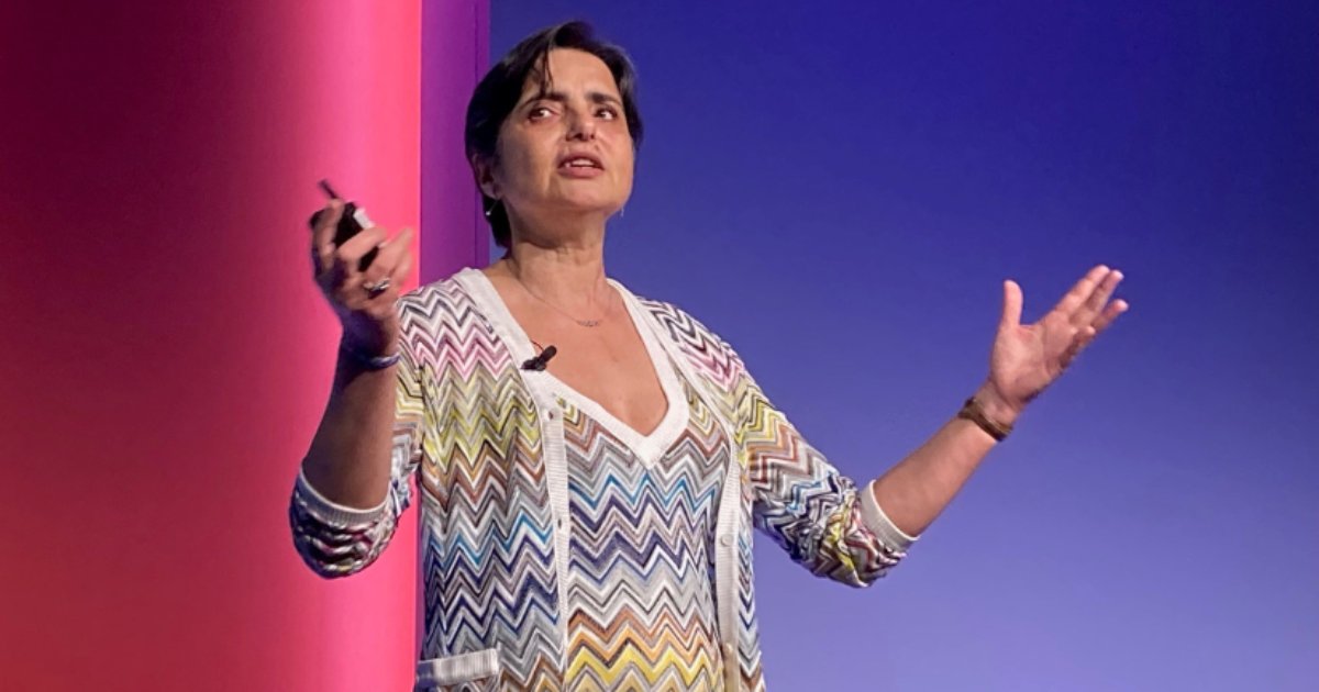 Roxane Edjlali, da Gartner, durante uma apresentação em um evento. Ela está em pé no palco, gesticulando com as mãos enquanto fala. Veste um cardigã colorido com padrão geométrico sobre uma blusa branca e usa um microfone de lapela. O fundo tem um degradê de cores entre azul e rosa, destacando a iluminação do evento (dados)