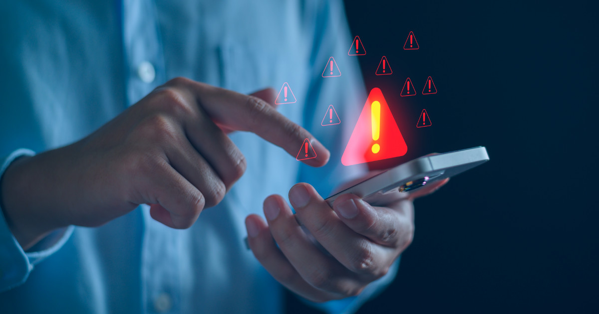 A imagem mostra uma pessoa segurando um smartphone enquanto toca na tela com o outro dedo. Diversos ícones vermelhos de alerta com pontos de exclamação aparecem sobrepostos à cena, simbolizando um aviso ou risco relacionado ao uso do dispositivo. O fundo escuro e desfocado destaca a interação e transmite uma sensação de preocupação ou atenção necessária, sugerindo um possível problema de segurança digital, golpe ou notificação de erro (avisamos)
