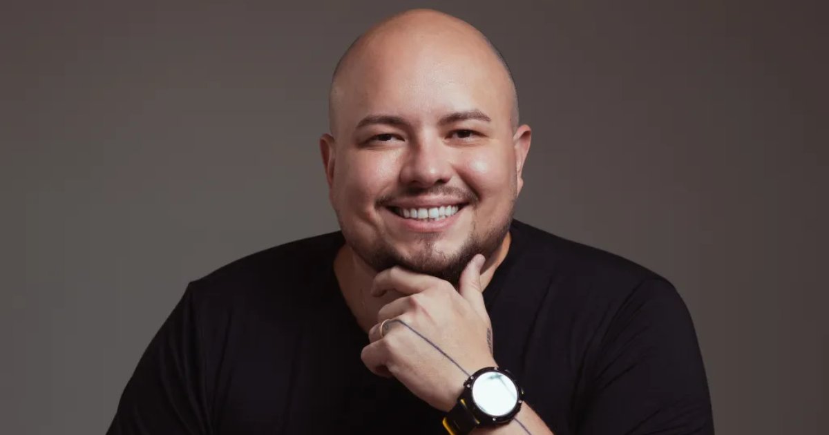 Fred Amaral, CEO da Lerian, em um retrato profissional. Ele é um homem careca e de barba curta, sorrindo enquanto apoia o queixo na mão. Usa uma camiseta preta e um relógio de pulso preto com detalhes dourados. O fundo é neutro e escuro, destacando sua expressão amigável, Midaz