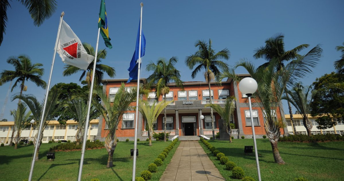 Prédio principal do Inatel (Instituto Nacional de Telecomunicações), com fachada de tijolos aparentes e janelas simétricas, rodeado por palmeiras e jardins bem cuidados. Na frente, três mastros com as bandeiras de Minas Gerais, do Brasil e do Inatel. Céu azul e clima ensolarado completam a cena (inatel, telecomunicações, telecom, minas gerais, mg)