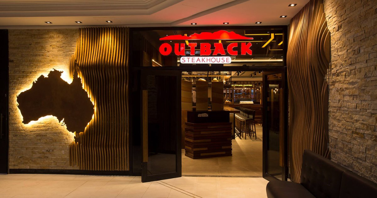 Fachada de uma unidade do restaurante Outback Steakhouse. A entrada possui iluminação aconchegante, com destaque para o letreiro vermelho e branco com o nome da marca e o contorno de uma paisagem desértica. Ao lado esquerdo da porta, há uma escultura iluminada no formato do mapa da Austrália. (outback stakehouse, nuvem)