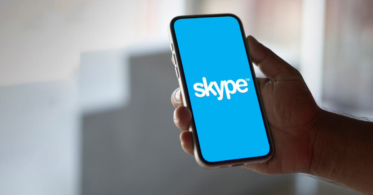A imagem mostra uma mão segurando um smartphone com a tela exibindo o logotipo do Skype em um fundo azul. O fundo da imagem está desfocado, sugerindo um ambiente interno com iluminação natural