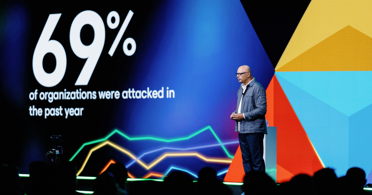 Um homem careca, de óculos e vestindo casaco cinzento e calças escuras, está em pé sobre um palco a fazer uma apresentação. Atrás dele, há um ecrã gigante com gráficos coloridos e texto em inglês que diz: “69% of organizations were attacked in the past year” (“69% das organizações foram atacadas no último ano”). A plateia está em silhueta no primeiro plano, observando atentamente. O cenário tem elementos gráficos coloridos em azul, vermelho, laranja e amarelo, criando um fundo moderno e tecnológico (Anand Eswaran, CEO da Veeam)