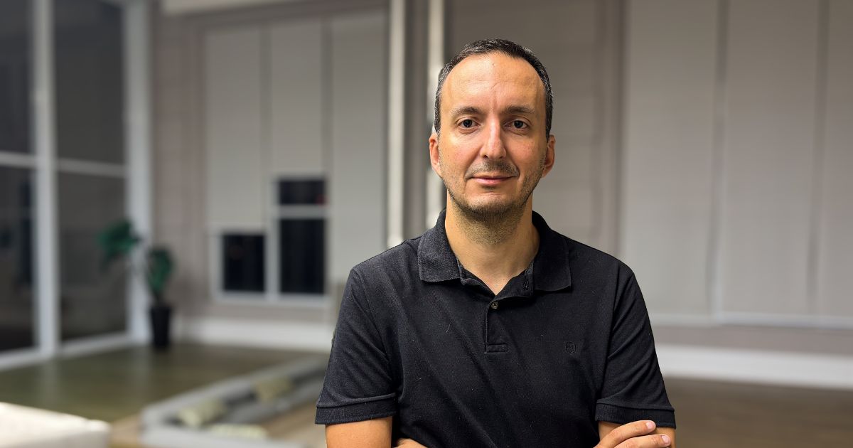 Bruno Picosse, diretor da Accenture Song, está de braços cruzados e veste uma camisa polo preta. Ele está em um ambiente interno moderno, com paredes de vidro, persianas fechadas e uma planta decorativa ao fundo. A iluminação é suave e o foco está no retrato profissional.