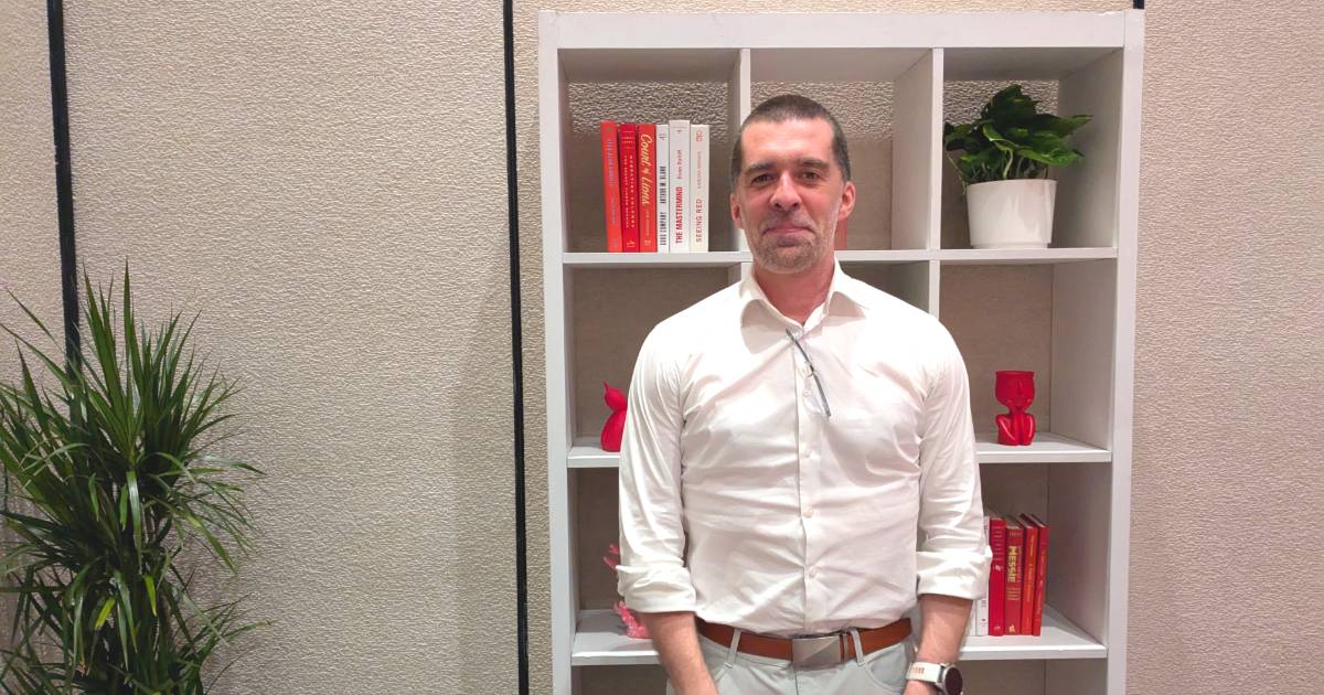 Homem em pé, usando camisa social branca e calça clara, com óculos pendurado na gola da camisa. Ao fundo, há uma estante branca com livros, objetos decorativos vermelhos e um vaso de planta. À esquerda da imagem, há uma planta decorativa. Trata-se de Mat Velloso, VP de Produto - Plataforma de Desenvolvimento em IA do Google DeepMind