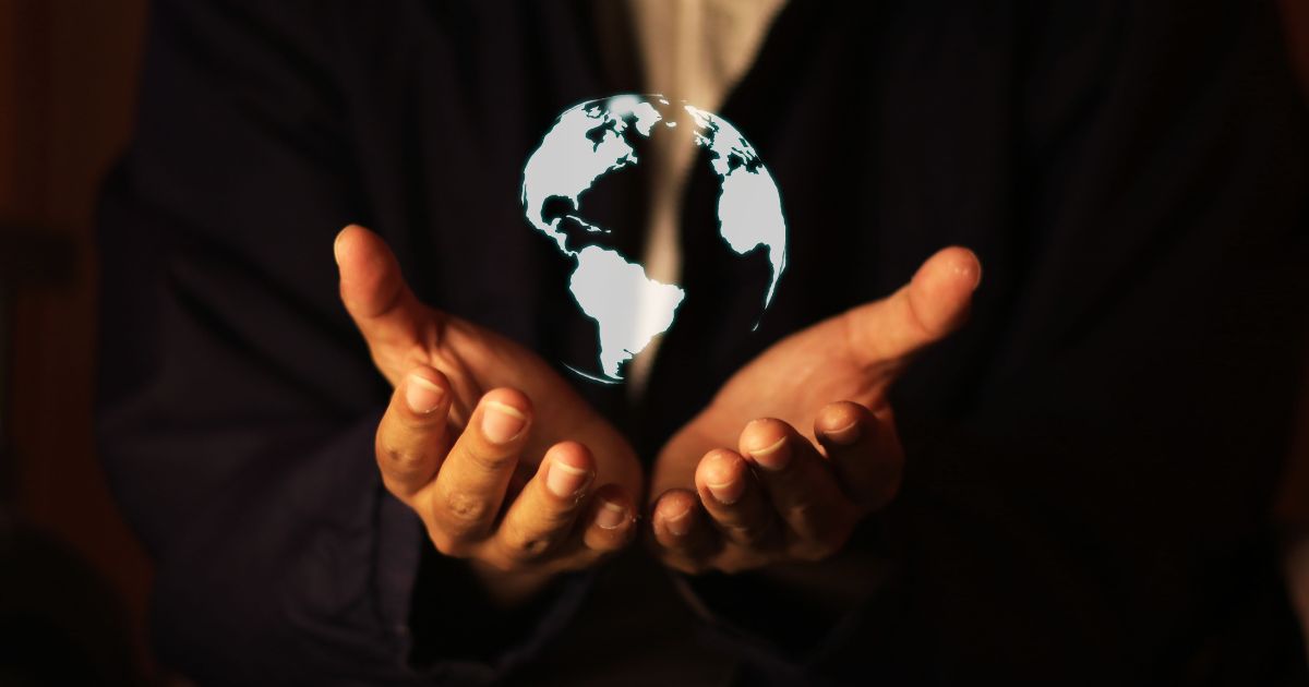 Uma pessoa, vestida com roupas escuras, estende as mãos abertas, com as palmas voltadas para cima. No centro, flutua uma representação digital e brilhante do planeta Terra, destacando os continentes iluminados em contraste com o fundo escuro. A imagem transmite uma sensação de poder, responsabilidade e conexão com o mundo (Axeptio)