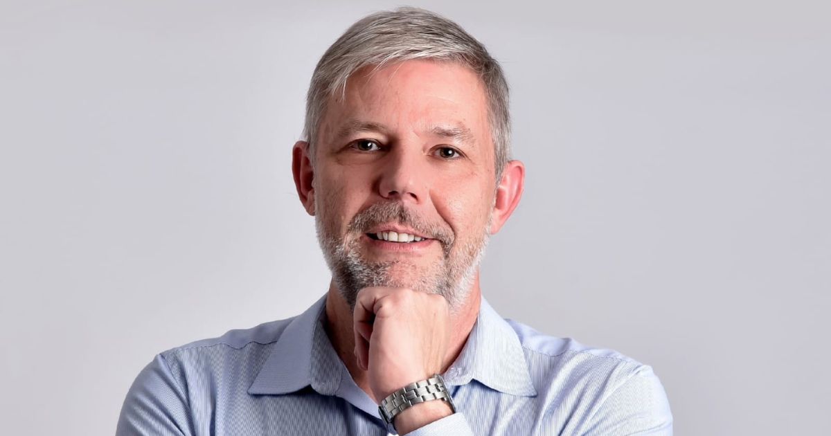 Homem de meia-idade com cabelo grisalho curto e barba por fazer, usa uma camisa azul clara e relógio de pulso metálico. Ele está a sorrir levemente, com a mão direita apoiada no queixo, num gesto pensativo. O fundo é simples e de cor clara, destacando a figura central. (Plano & Plano)