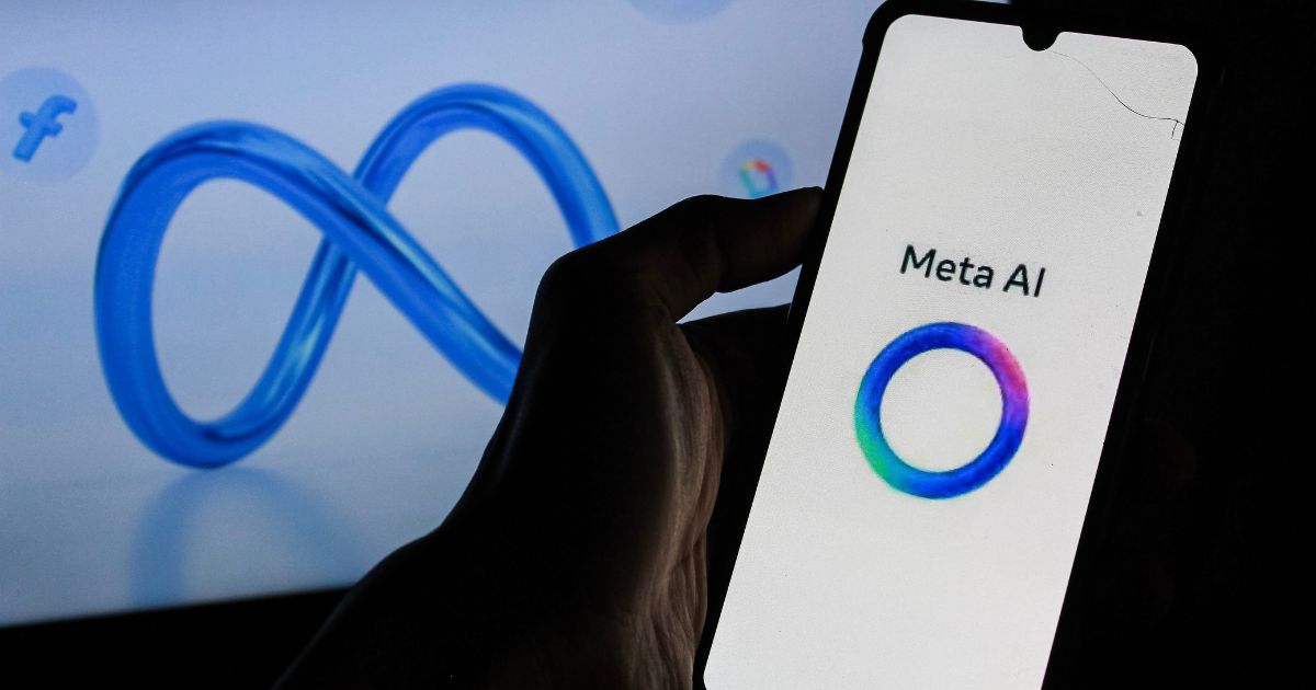 Fotografia de uma mão a segurar um smartphone que exibe o logótipo e o nome 'Meta AI' num ecrã branco. Ao fundo, desfocado, vê-se o logótipo azul em forma de infinito da empresa Meta, acompanhado de pequenos ícones das suas plataformas