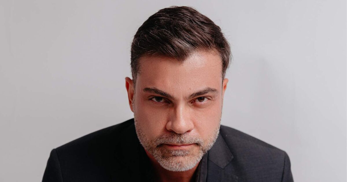 Retrato de Lúcio Oliveira, executivo de tecnologia e terapeuta. Ele está com expressão séria, usando terno escuro, olhando diretamente para a câmera, com fundo neutro. Plano B