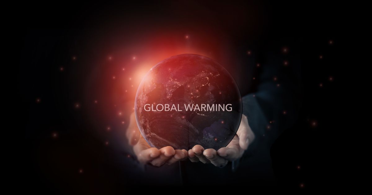 Imagem simbólica de duas mãos a segurar um globo terrestre iluminado por uma luz vermelha intensa, representando o aquecimento global. No centro do planeta, em destaque, aparece o texto 'GLOBAL WARMING'. O fundo é escuro, com pequenas partículas luminosas espalhadas, sugerindo calor ou partículas no espaço.