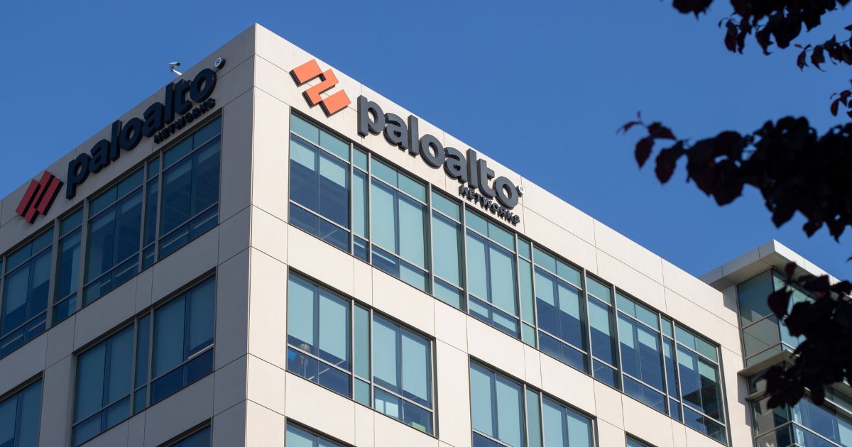 "Imagem de um prédio corporativo com fachada de vidro, onde está o logotipo da empresa Palo Alto Networks no topo. A marca, conhecida por atuar na área de cibersegurança, aparece em letras pretas ao lado de um símbolo gráfico vermelho. O céu azul e claro ao fundo reforça o ambiente empresarial moderno."