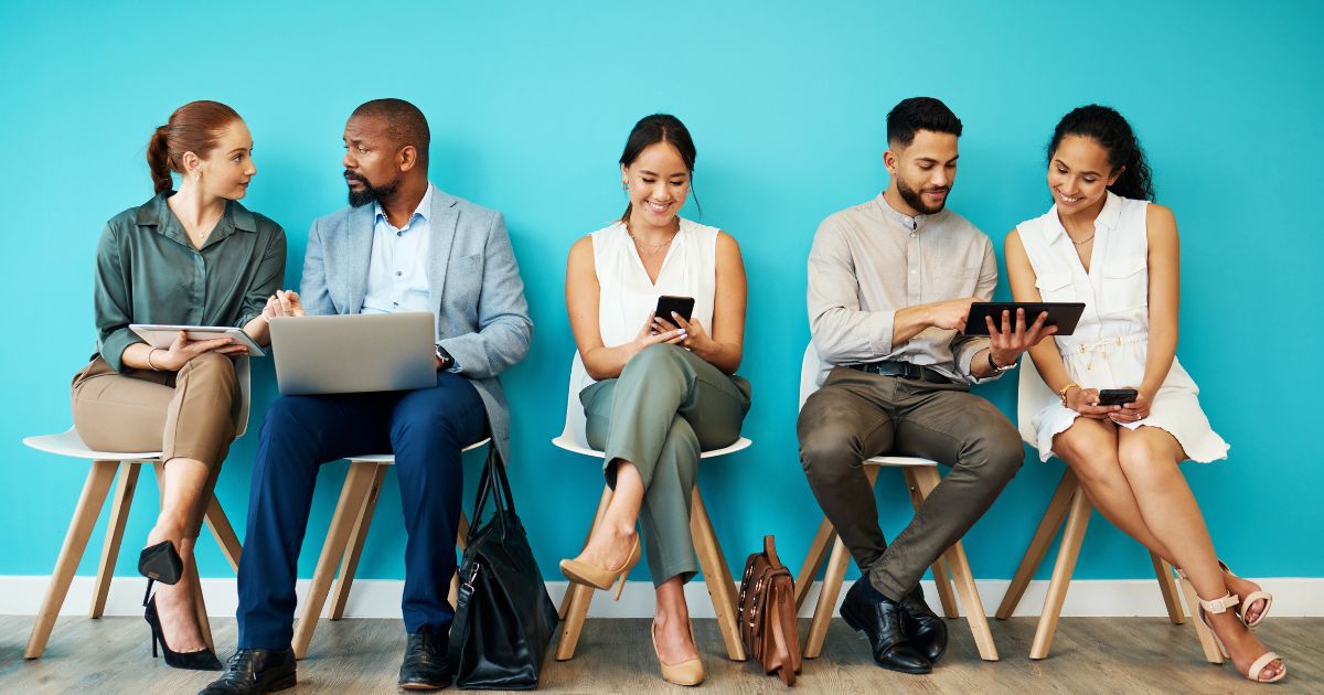Seis pessoas sentadas lado a lado em cadeiras brancas, frente a uma parede azul. Estão todas bem vestidas, em ambiente informal-profissional, usando dispositivos como portáteis, tablets e telemóveis. Estão envolvidas em conversas ou concentradas nos seus ecrãs, transmitindo diversidade e colaboração num ambiente de trabalho moderno.