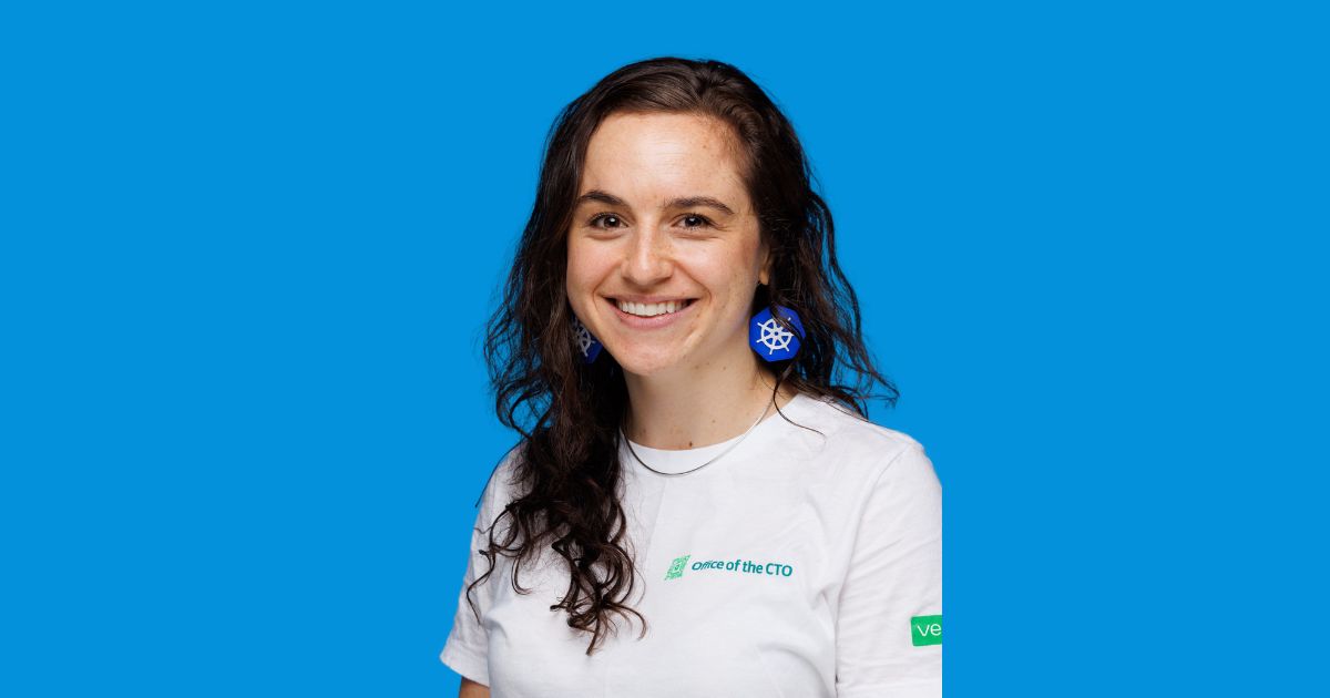 Uma mulher jovem com cabelo castanho-escuro e ondulado sorri para a câmara, em frente a um fundo azul vivo. Ela está vestida com uma t-shirt branca onde se lê “Office of the CTO” e exibe brincos grandes em forma do logótipo do Kubernetes (roda de leme azul). Ela transmite uma imagem descontraída e simpática, num contexto claramente profissional ligado à tecnologia (kubernetes)