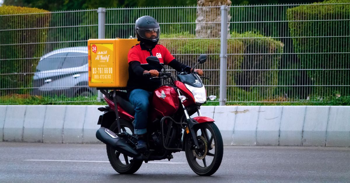 Um motociclista usando capacete preto, jaqueta vermelha e preta, calça jeans e botas está pilotando uma moto vermelha em uma avenida. Na parte traseira da moto, há uma grande caixa amarela com inscrições em árabe e inglês, indicando que se trata de um serviço de entrega. O fundo mostra uma cerca metálica, arbustos podados e um carro cinza estacionado ao lado da via.