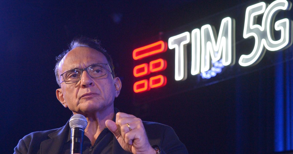 Homem de meia-idade, com óculos e expressão séria, segura um microfone enquanto fala em público. Ele está vestido com um casaco escuro e usa uma aliança dourada na mão esquerda. Ao fundo, vê-se um letreiro iluminado com as palavras "TIM 5G" e o logótipo da empresa TIM, destacando-se num ambiente escuro com luzes azuis.
